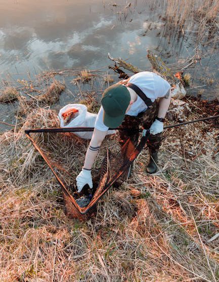 A dedicated volunteer uses a net to clean trash by a serene riverside during sunset.