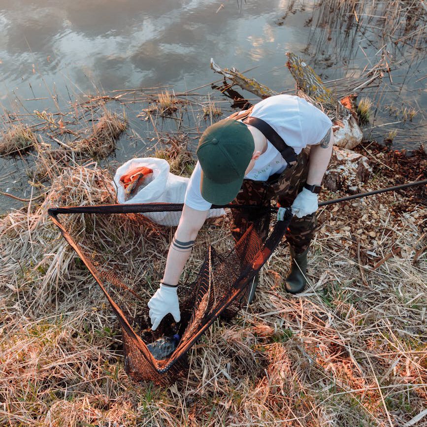 A dedicated volunteer uses a net to clean trash by a serene riverside during sunset.
