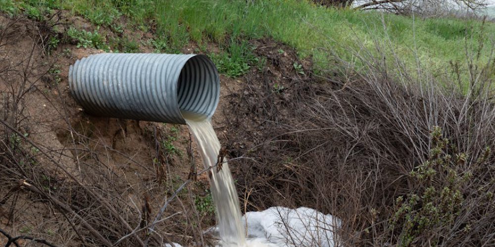A rural drainage pipe pouring water into a natural ravine, surrounded by green vegetation.