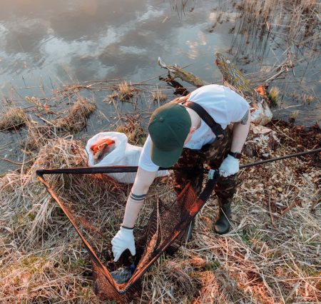 A dedicated volunteer uses a net to clean trash by a serene riverside during sunset.